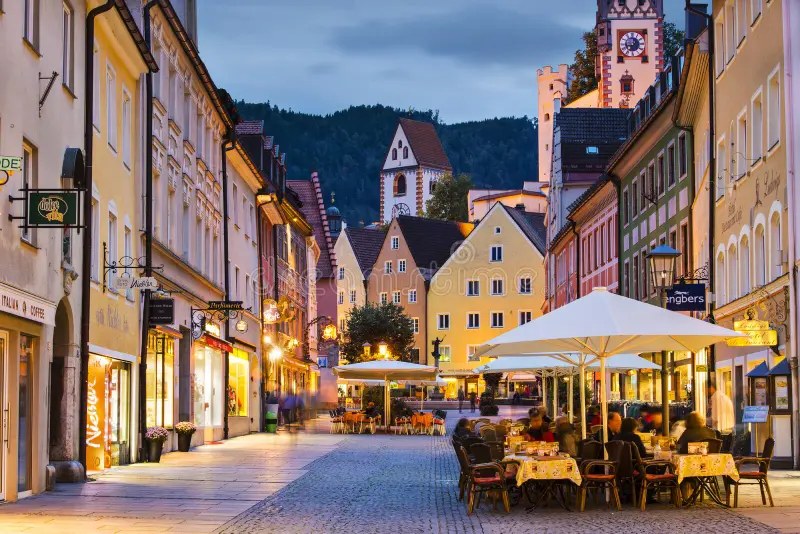 Abendliche Fußgängerzone in der Füssener Altstadt mit bunten Häusern, Cafétischen und dem Uhrturm des Hohen Schlosses