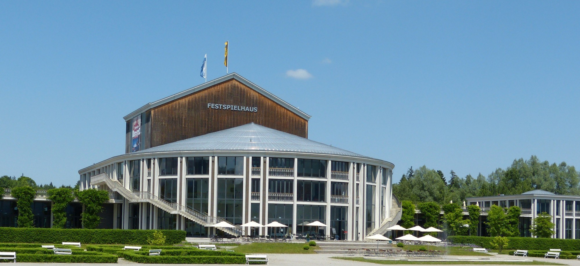 Das Festspielhaus Füssen mit runder Glasfassade und Gartenanlage unter blauem Himmel