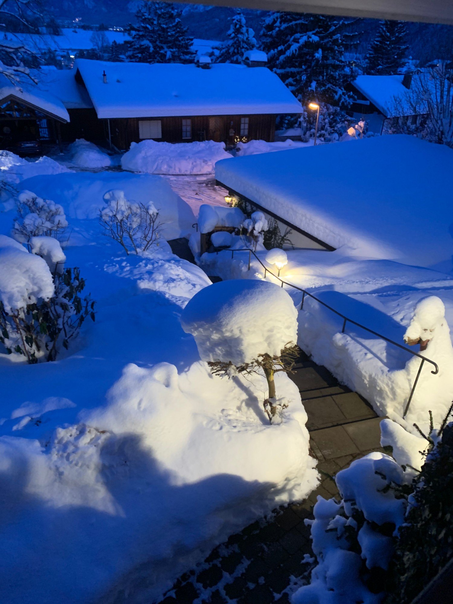 Verschneite Nachbarschaft am Abend mit Laternenlicht, tief verschneiten Büschen und Holzhäusern